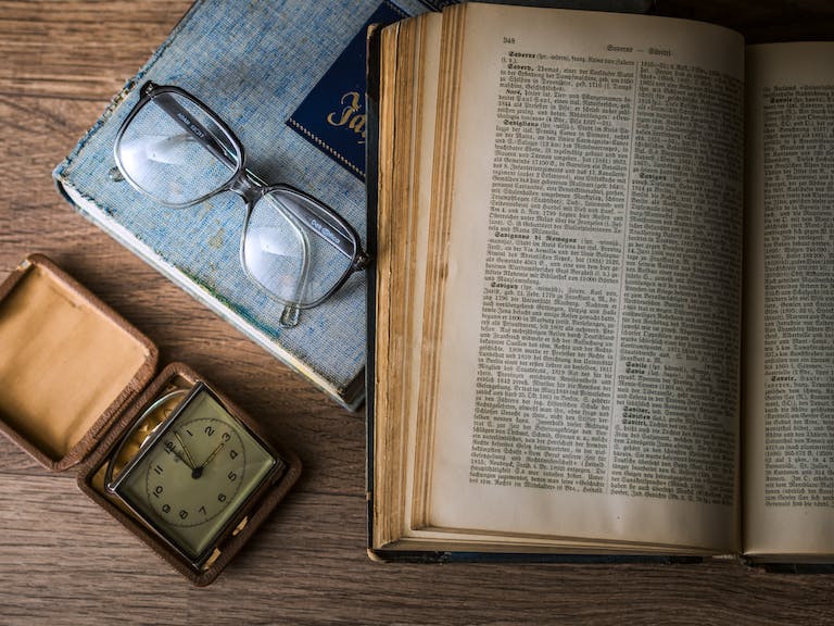 Opened Book Beside Sunglasses and Square Gray Clock Displaying 3:00 Time