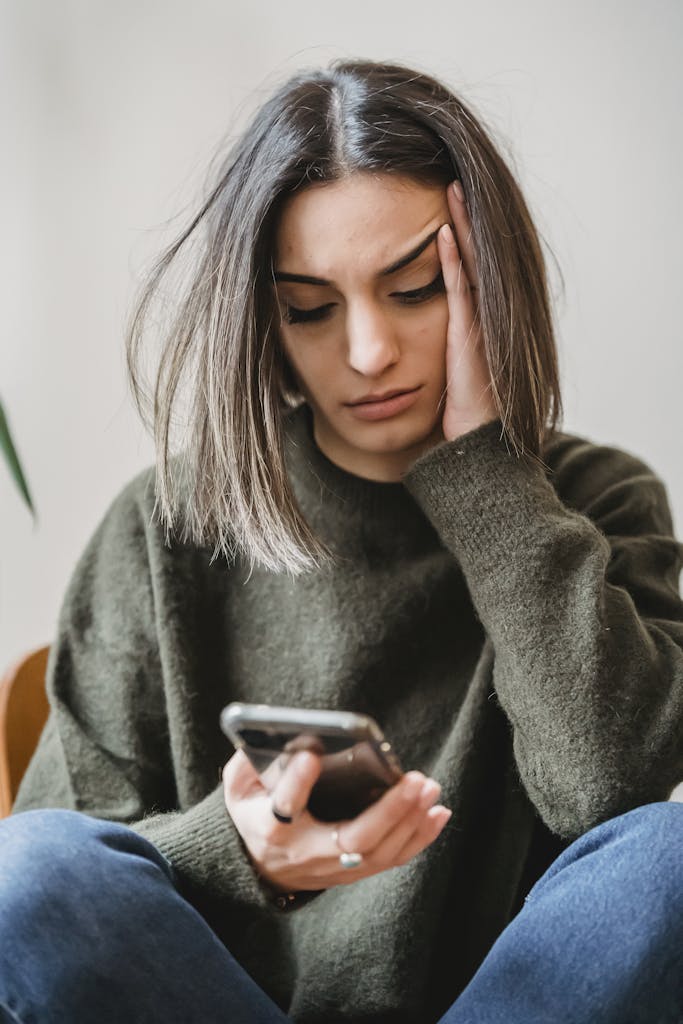 Unhappy female in casual wear leaning on hand and surfing internet on cellphone while sitting in light room near wall at home