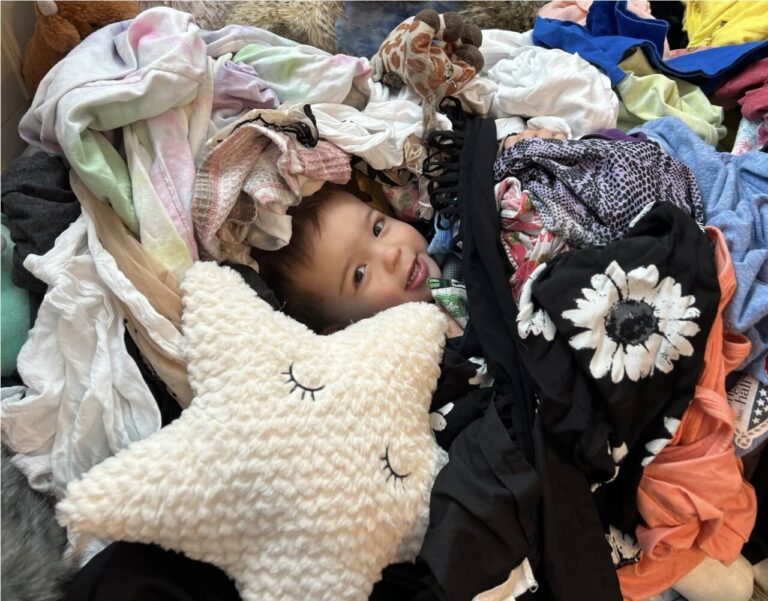 a baby hiding among a pile of clothing and toys.