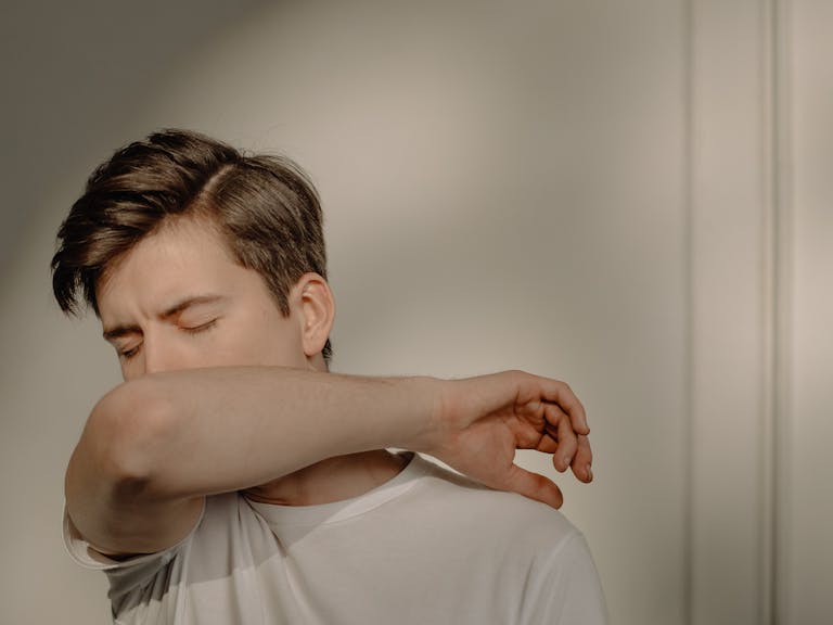 A young man sneezing into his elbow indoors, emphasizing health and hygiene.