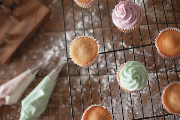 Top view of colorful frosted cupcakes on a cooling rack with piping bags and flour dust.