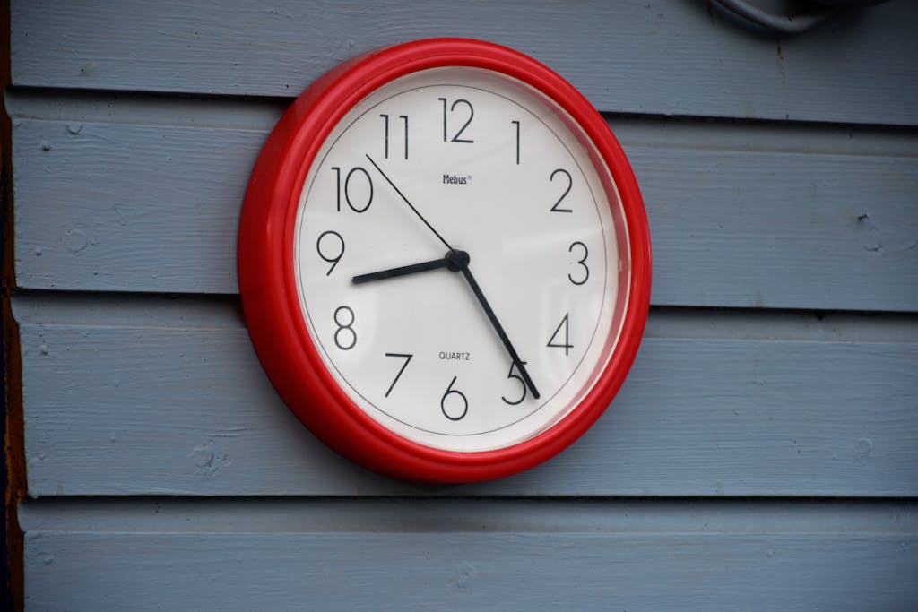A red wall clock with black hands on a blue wooden wall, captured in daylight.