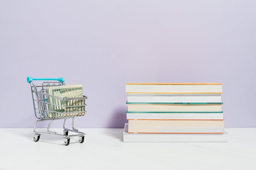 A shopping cart with dollar bills next to a stack of textbooks on a light background.