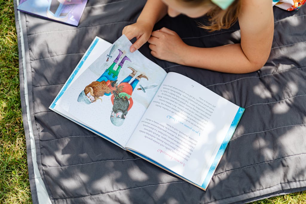 A young girl enjoys reading on a blanket in the sunlit outdoors, perfect summer relaxation.