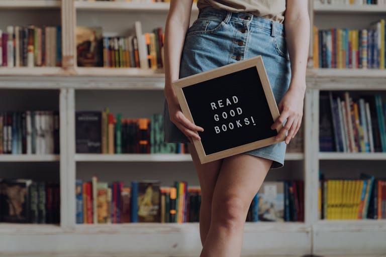 Person holding a sign with 'Read Good Books!' in a bookstore setting, encouraging reading culture.