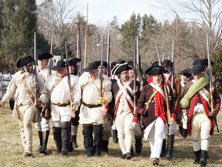 Revolutionary War reenactors in period uniforms march together outdoors in Washington Crossing, PA.