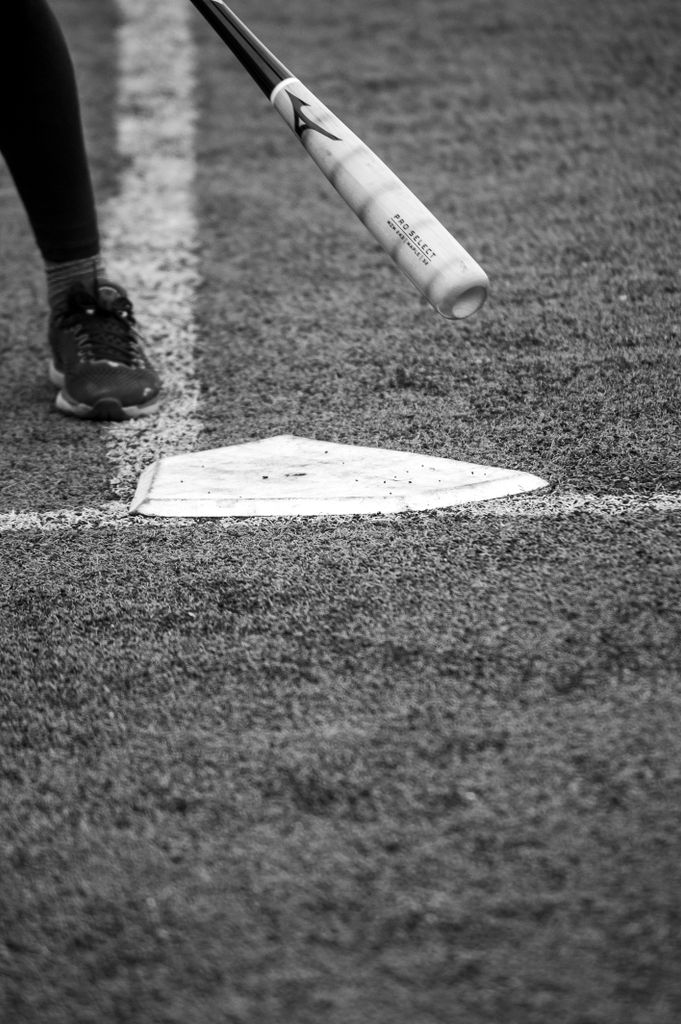 Monochrome close-up of a baseball bat near home plate on a field with player foot visible.
